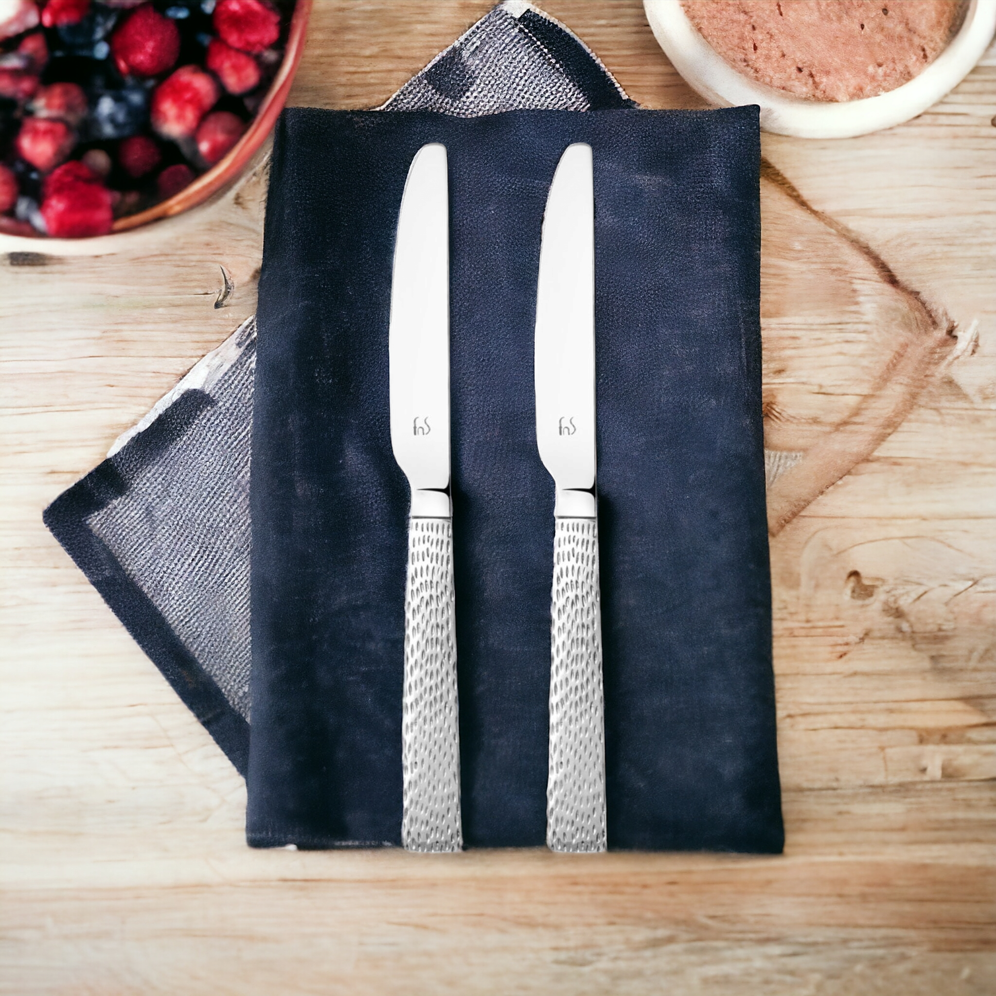 Pair of stainless steel dinner knives with decorative mesh-pattern handles on black slate board with berries and coffee on wooden table.