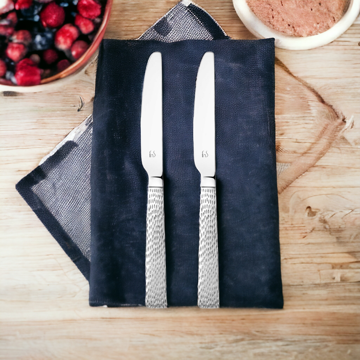 Pair of stainless steel dinner knives with decorative mesh-pattern handles on black slate board with berries and coffee on wooden table.