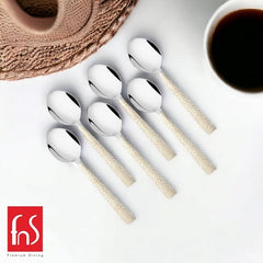 Six stainless steel tea spoons with textured beige handles arranged on white table with coffee cup and woven placemat