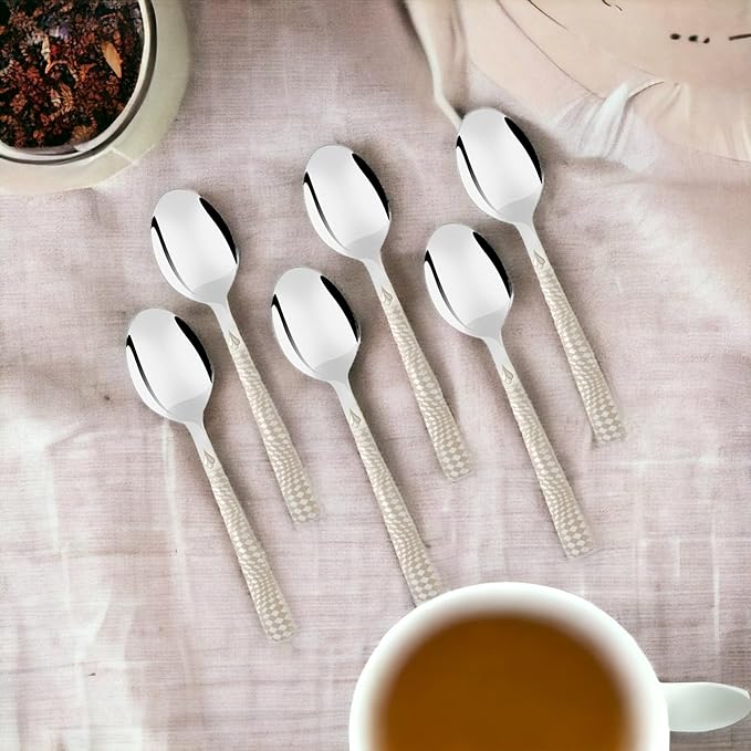 Six stainless steel tea spoons with textured white handles arranged on beige linen fabric with tea cup and dessert bowl