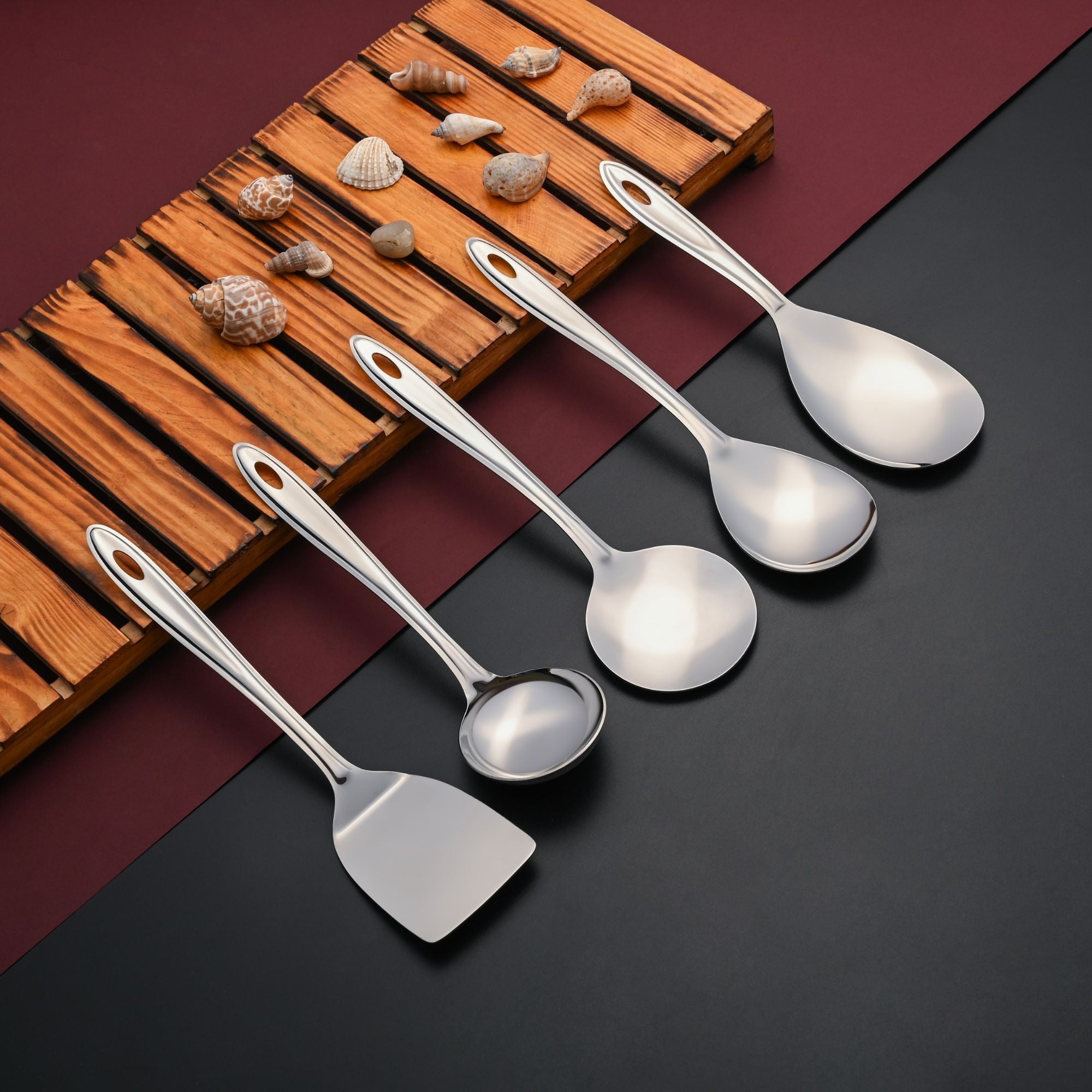 Trinity serving tools including spoons, ladle, and spatula arranged on wooden board with seashells and burgundy accent
