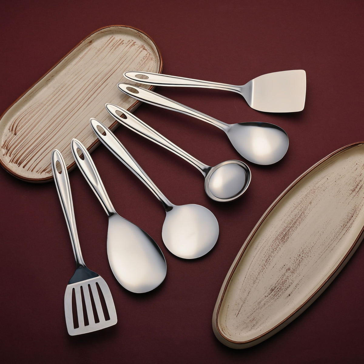 Trinity 6-piece serving tools set arranged on ceramic plates, featuring polished stainless steel spatulas, ladles, and spoons for complete kitchen serving.