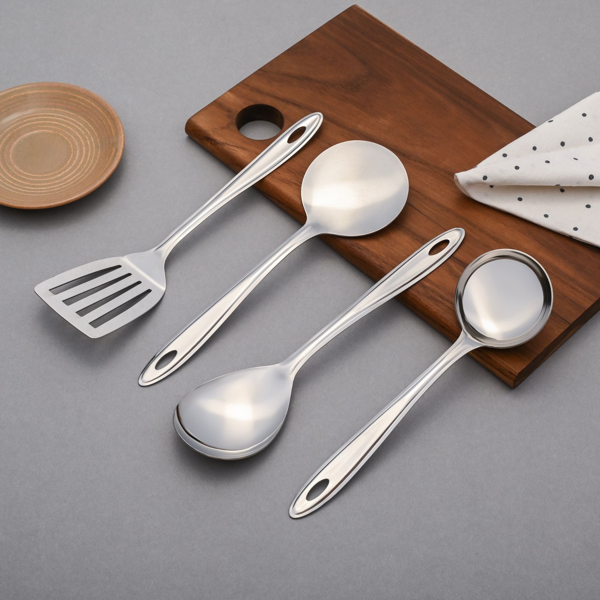Trinity serving tools including slotted turner, ladle, and spoons displayed on wooden cutting board with ceramic plate