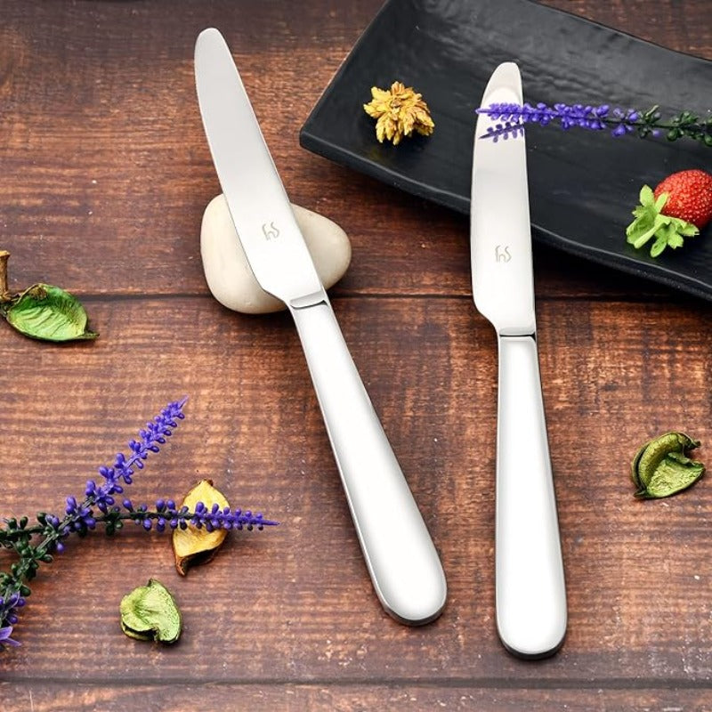 Two stainless steel butter knives on rustic wooden table with purple lavender, strawberry, golden leaf, and black serving plate creating elegant flatware display.
