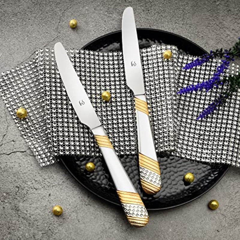 Two stainless steel dinner knives with gold and silver decorative handles on black plate with houndstooth napkin and purple flowers.