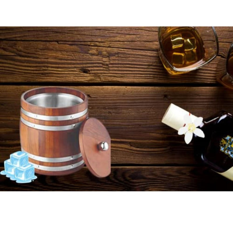 Wooden barrel-style ice bucket with stainless steel bands and wooden lid on rustic wooden table with ice cubes, whiskey glass, candle, and white orchid