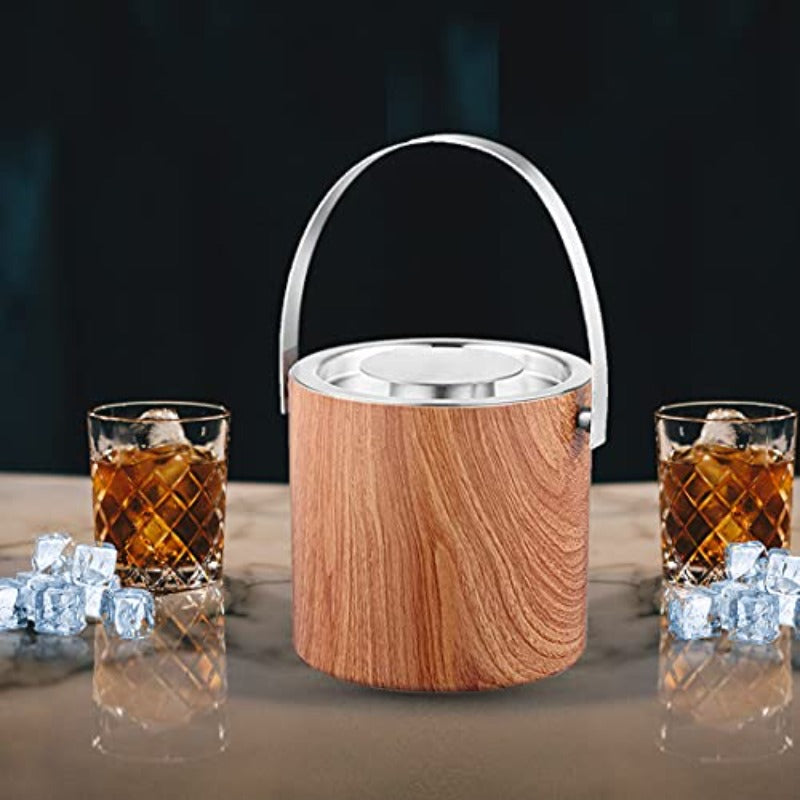 Wooden finish ice bucket with stainless steel rim and handle displayed with two crystal whiskey glasses filled with ice and amber liquid on marble counter