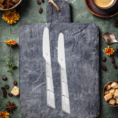 Styled flat lay of two silver dinner knives on dark slate board surrounded by decorative spices, yellow flowers, coffee cup, sugar cubes, and scattered coffee beans on textured green surface.